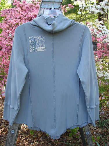 Barclay Patched Thermal Cowl Neck Roo Tunic Gray Day Size 1, featuring raw seams, a pointed hemline, exposed stitchery, and a distinctive chest patch, displayed outdoors on a wooden rack.