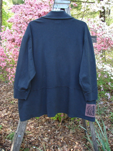 2000 Interlock Peat Bog Top Unpainted Black Size 2 displayed outdoors on a wooden support, showcasing its deep V-shaped neckline, unique kangaroo pocket, and signature Blue Fish patch against a garden backdrop.