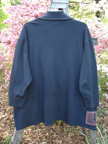Back view of the 2000 Interlock Peat Bog Top Unpainted Black Size 2, featuring a deep V-neck and kangaroo pocket, hanging outdoors against a backdrop of pink flowering shrubs.