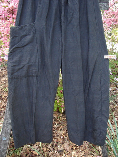 Barclay Linen Striation Single Side Pocket Pant Unpainted Black Generous Size 0, gracefully draped on an outdoor rail, showcases its loose-fit, wide-leg design with a prominent side patch pocket, framed by pink flowering shrubs.