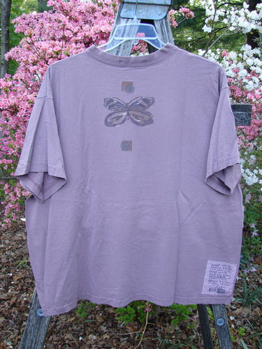1996 Short Sleeved Tee Triple Butterfly Petal Size 2 displayed on a hanger outdoors, showcasing its stylized butterfly motif and signature patch, set against a backdrop of blooming shrubs.