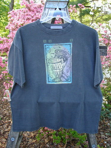1996 Short Sleeved Tee Drummer Storm Size 1, featuring a unique abstract graphic of trash cans with swirling accents. Displayed on a hanger outdoors against flowering shrubs, highlighting its vintage Blue Fish Clothing style.