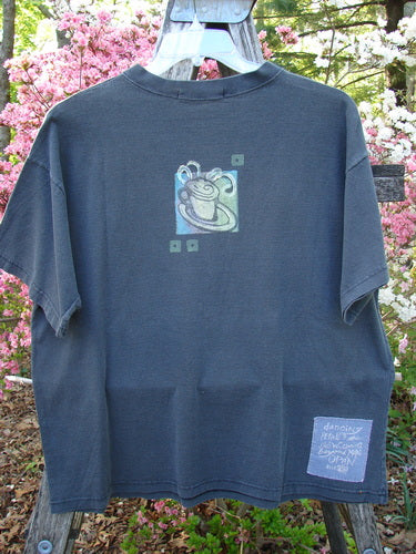 1996 Short Sleeved Tee Drummer Storm Size 1 displayed on a wooden ladder, featuring a central steaming coffee cup motif and a signature patch. Set against a backdrop of flowering shrubs and foliage.