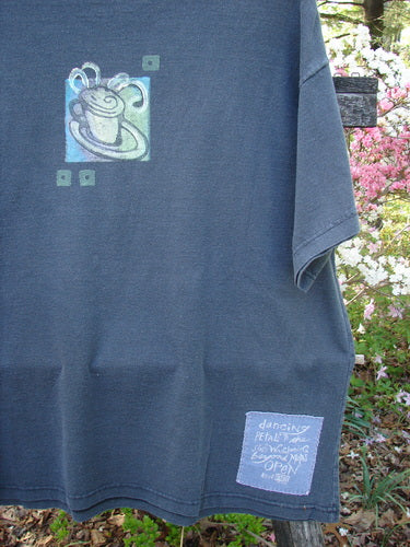 1996 Short Sleeved Tee Drummer Storm Size 1 features a swirly flower motif and text patch, hung outdoors among flowering shrubs, showcasing Bluefishfinder.com's unique vintage style.