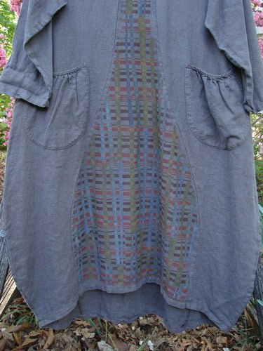 Barclay Linen Tea Ceremony Thostle Dress Cross Grid Dark Stone Size 1, featuring ¾-length sleeves, front-seam patch pockets, and a central plaid panel, hanging outdoors against lush foliage.