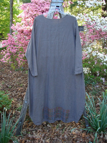 Barclay Linen Tea Ceremony Thostle Dress Cross Grid Dark Stone Size 1 hangs outdoors, showcasing its loose fit, long sleeves, and subtle hem detail against a backdrop of flowering shrubs.