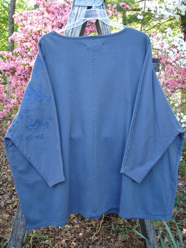 Barclay Fleece Big Square Pullover Botanical Path Night Navy OSFA hangs on a ladder, showcasing dolman shoulders, narrowing sleeves, and subtle floral embroidery, set against a vibrant garden backdrop.