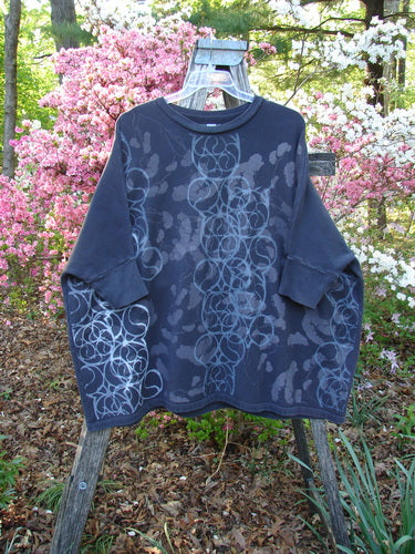 Barclay Fleece Banded Sleeve Dolman Top features a wide, boxy shape with continuous spirograph patterns. Displayed outdoors against flowering shrubs, it embodies Bluefishfinder's unique vintage style in cozy, heavyweight organic cotton fleece.