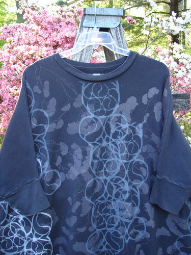 Barclay Fleece Banded Sleeve Dolman Top, featuring overlapping light-grey rings on dark-charcoal fabric, hangs on a plastic hanger. Showcasing the cozy, artistic style of Bluefishfinder.com, perfect for fall.