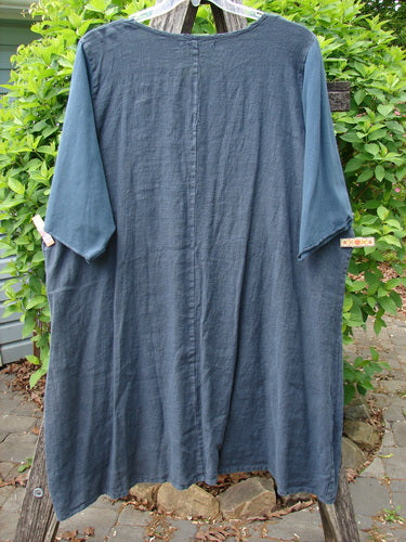 Barclay NWT Linen Cotton Sleeve Milkmaid Dress in Blue Cypress, featuring sunrise pleats, empire waist, and diagonal pockets. Hangs on a wooden frame with greenery backdrop, showcasing its unique silhouette.