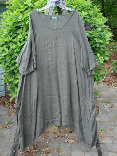 Barclay Linen Drop Pocket Bell Dress Unpainted Stick Size 2 hangs on a hanger, featuring elbow-length sleeves, scooped neckline, large pockets, and an A-line silhouette. Photographed outdoors against greenery.