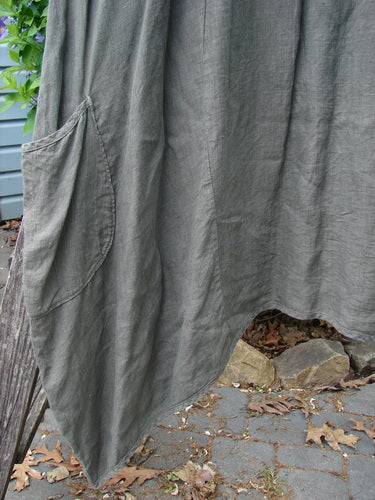 Barclay Linen Drop Pocket Bell Dress Unpainted Stick Size 2 features an asymmetrical handkerchief hem and curved patch pocket, showcasing textured medium-weight linen hanging over stone-and-leaf ground.