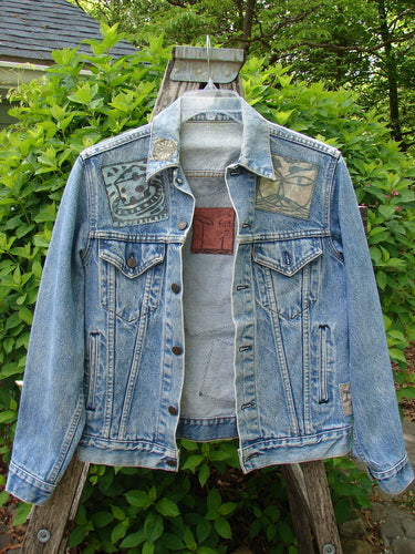 1995 Patched Levi Strauss & Co Denim Blue Jean Jacket Kitty Kitty Stone Wash Size 0 displayed on a rustic ladder, featuring chest flap pockets, abstract shoulder patches, and a stand-up collar amidst a leafy backdrop.