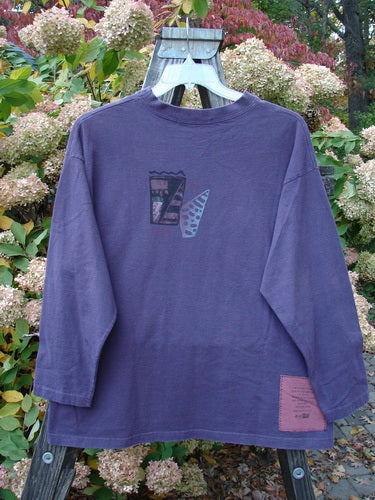 A long sleeved purple shirt featuring a thicker mid weight cotton, wider ribbed neckline, and cozy longer sleeves. The shirt is adorned with an intriguing Spirit Woman and Drum theme paint. Size 1.