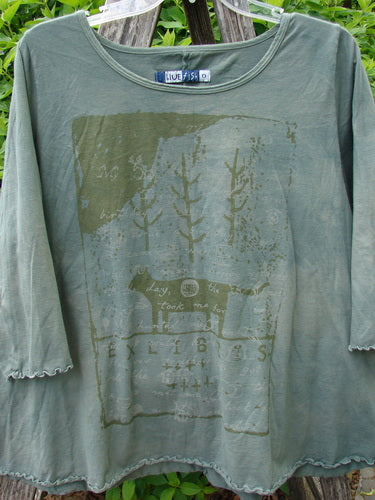 Barclay Crop A Lined Cafe Tee Cow Farm Grass Size 0: Loose-fitting olive-green top with stylized tree and animal graphic, curly edgings, and a rounded neckline, hanging in front of foliage.