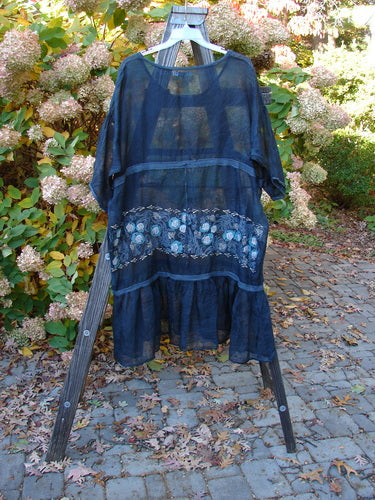 A blue dress with flowers on a wooden rack, featuring a rounded neckline, empire waist seam, and full drawcord gather front. The dress has a sectional lower, silken edgings, and horizontal banded accents. It also has a complete full ruffled lower and generous garden patch theme paint. Size 2.