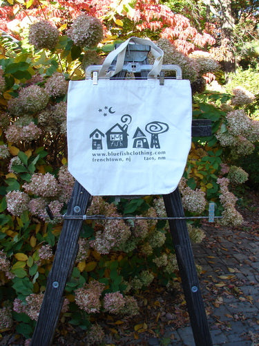 Image: A white bag with black text on it, placed on a ladder. The bag is made of lighter weight cotton canvas and has a tightly woven shorter shoulder strap. The bag features a generous top opening and a deeper rounded bottom. The text on the bag celebrates the Blue Fish Barclay Shops and features the Village Logo. This Barclay Blue Fish Promo Tote Bag is from a Limited Edition Release and is in Perfect One Size Condition. The measurements are as follows: Strap 8, Width 18, Length 15 inches.