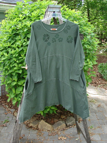 Barclay Cotton Lycra Shape Dress Wind Flower Deep Moss Size 1, featuring raw-edge sleeves, stenciled floral motifs, an A-line cut, and side pockets, displayed on a wooden stepladder amidst a garden setting.