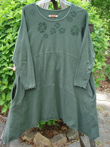 Barclay Cotton Lycra Shape Dress Wind Flower Deep Moss Size 1 features a forest-green A-line silhouette with scalloped hem, long sleeves, and flower accents near the neckline, hanging against shrubbery.