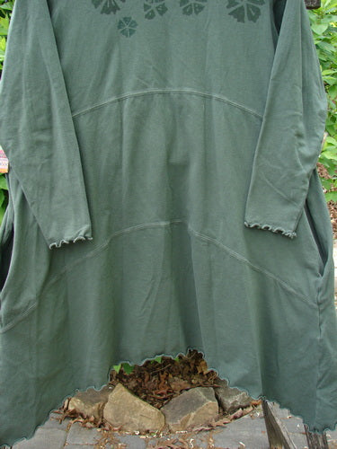 Barclay Cotton Lycra Shape Dress Wind Flower Deep Moss Size 1, featuring long sleeves, a scalloped hem, front patch pockets, and floral print on the chest, hangs outdoors against a leafy backdrop.