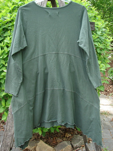 Barclay Cotton Lycra Shape Dress Wind Flower Deep Moss Size 1 features a flowing A-line with raw-edged seams, cozy long sleeves, side pockets, and an empire waist, hanging elegantly on a wooden ladder.