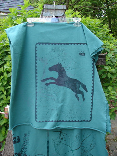 Barclay Heavy Weight Organic Sweatshirt Blanket Pony Garden Emerald OSFA displayed outdoors, featuring a black leaping horse silhouette within a stamp-like frame. The blanket has raw curly edges, embodying Blue Fish's creative style.