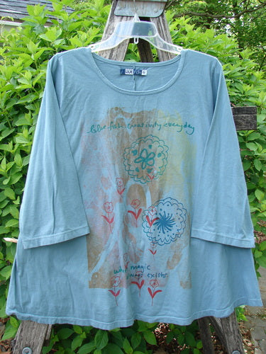 Barclay Three Quarter Sleeved A Lined Tee Creativity Everyday Dusty Sky Size 0 features a teal top with floral mandala designs and inspirational text, draped on a wooden ladder outdoors.