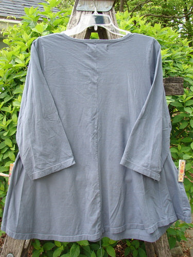 Barclay Three Quarter Sleeved A Lined Tee Wandering Dust Plum Size 0, showcases a slate-gray tunic-style top with a rounded neckline and curly edges, draped over a wooden ladder amidst greenery.