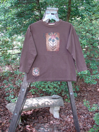 1996 Long Sleeved Tee Single Flower Molasses Size 0 displayed on a wooden ladder, featuring a thicker ribbed neckline, drop shoulders, longer square shape, and a painted single flower design.