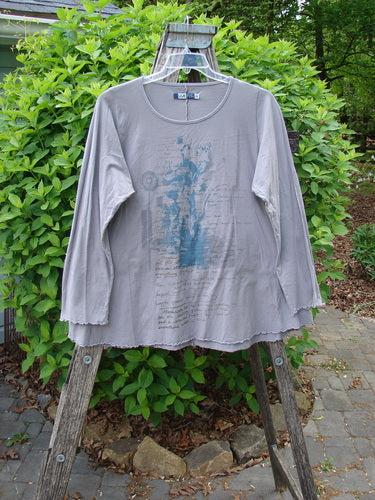 Barclay Long Sleeved Cafe Tee Poetry Garden Grey Day Size 0, featuring a rounded neckline and curly edgings, hangs on a ladder, showcasing a faded graphic amid a garden-themed backdrop.