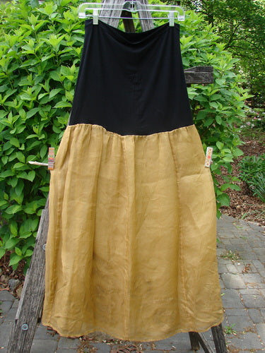 The 2000 Silk Organza Aios Dana Skirt Celtic Wind Bone Size 0, features a black upper and golden lower flare, elegantly displayed on a hanger against leafy shrubs, embodying Bluefishfinder.com's vintage charm.