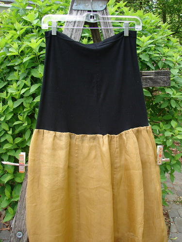 2000 Silk Organza Aios Dana Skirt Celtic Wind Bone Size 0 features a solid black top with a gathered golden-tan bottom, elegantly displayed on a hanger against leafy shrubs.