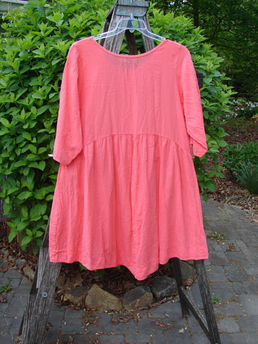 Barclay Linen Two Pocket Dolly Dress Notes Coral Size 0 hangs on a clear hanger, showcasing its flouncy lower skirt, wide hem sweep, and dual exterior drop pockets against a natural outdoor backdrop.