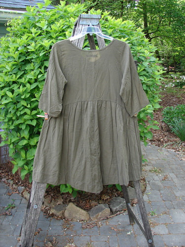Barclay Linen Two Pocket Dolly Dress with elbow-length sleeves and a flouncy gathered skirt, showcasing drop pockets and brush paint design, elegantly hangs on a wooden ladder outdoors.