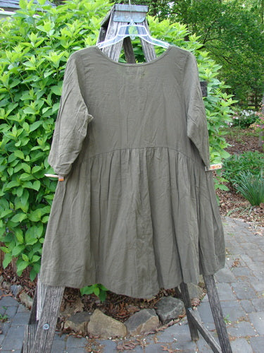 Barclay Linen Two Pocket Dolly Dress Brush Paint Wood Size 0 draped over a wooden ladder outdoors, showcasing its flouncy lower skirt, dual drop pockets, and wide hem sweep in a garden setting.