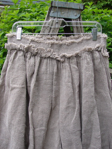 Magnolia Pearl Homespun Linen Crop Smocked Bloomer Flaxen OSFA, featuring a gathered skirt with a frayed-edge waistband, hangs on a wooden ladder, showcasing its textured, natural linen fabric and signature patches.