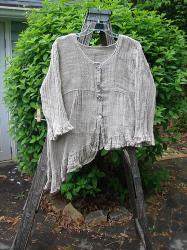 Magnolia Pearl Fisherman's Weave Asymmetrical Ruffle Jacket draped over a wooden A-frame, showcasing its ruffled hem, oversized pearl buttons, and a unique downward curved bodice against a backdrop of green shrubs.