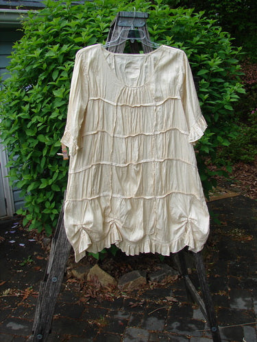 Magnolia Pearl Silk Cotton Drapery Dress Butter Creme OSFA, featuring a scoop neckline, elbow-length sleeves, and layered ruffled tiers, displayed on a wooden ladder against lush greenery.
