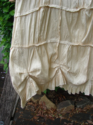 Magnolia Pearl Silk Cotton Drapery Dress Butter Creme OSFA, elegantly draped over a wooden railing, showcases its tiered, ruffled design with gathered seams and a full ruffled hemline, embodying vintage charm.