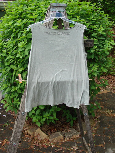Magnolia Pearl Cotton Jersey Vented Layer Tank Top Green River OSFA draped on a clear hanger, featuring metallic-studded neckline, raw flutter edges, and distressed detailing against a rustic wooden ladder backdrop.