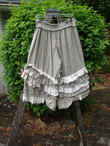 Magnolia Pearl Linen Eiffel Tower Bloomer Flaxen OSFA draped over a wooden easel, showcasing its ruffled tiers and smocked waist, set against lush green shrubs, embodying vintage elegance.