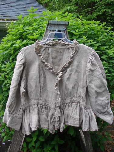 Magnolia Pearl Homespun Linen Snap Jacket Flaxen OSFA, features a ruffled neckline and snap line, peplum hem, and signature pearl patch, elegantly displayed on a hanger against lush greenery.