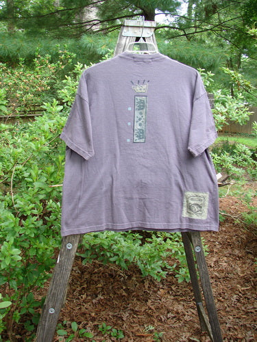 Alt text: 1995 Short Sleeved Tee Silly Pup Bloomsberry Altered Size 2 displayed on a wooden hanger outdoors, showcasing its thick ribbed neckline and detailed flower vase with butterflies design.