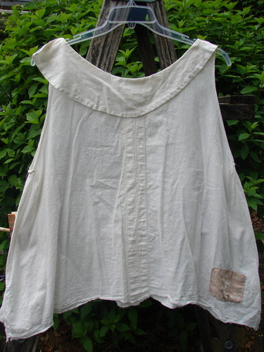 Magnolia Pearl Cotton Voile Sweet Collar Top White OSFA: An off-white, sleeveless garment with a wide collar, center seam, and signature love patch, displayed outdoors against leafy shrubs.