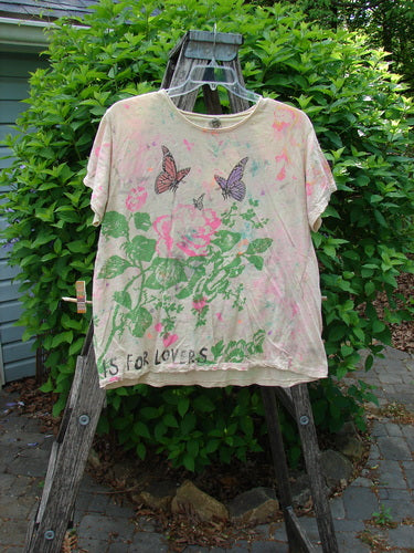Magnolia Pearl Cotton Jersey Boyfriend Tee features a pink rose, green foliage, and butterflies, casually displayed on an outdoor ladder, embodying Bluefishfinder.com's vintage and expressive style.