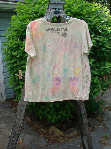 Magnolia Pearl Cotton Jersey Boyfriend Tee draped over a stepladder, featuring vibrant paint splatters, MAGNOLIA PEARL text on the back, and a lightweight, soft slub jersey fabric with distressed edges.
