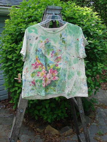 Magnolia Pearl Cotton Jersey Boyfriend Tee Songbird Floral OSFA displayed on a wooden stand, features a songbird print amid pink blossoms. Its lightweight, drapey cotton jersey shows unique shoulder distressing.