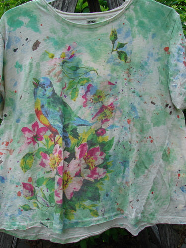 Magnolia Pearl Cotton Jersey Boyfriend Tee Songbird Floral OSFA features a vibrant watercolor bird amid pink blossoms on a light-green backdrop, draped over an outdoor wooden rail, showcasing its soft, drapey cotton fabric.