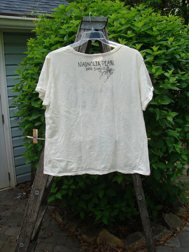 Magnolia Pearl Cotton Jersey Boyfriend Tee Iris Garden OSFA displayed outdoors on a wooden ladder. Features include a loose fit, drapey feel, and rear signature stamp with Live Slowly, Love Deeply text.