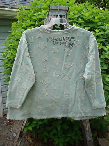 Magnolia Pearl Fleece Applique Little Star Hermosa Sweatshirt in Garden Sage hangs on a wooden ladder, showcasing raw-edged star appliqués, frayed hem, and printed “MAGNOLIA PEARL LOVE SUPPLY CO.” above the stars.